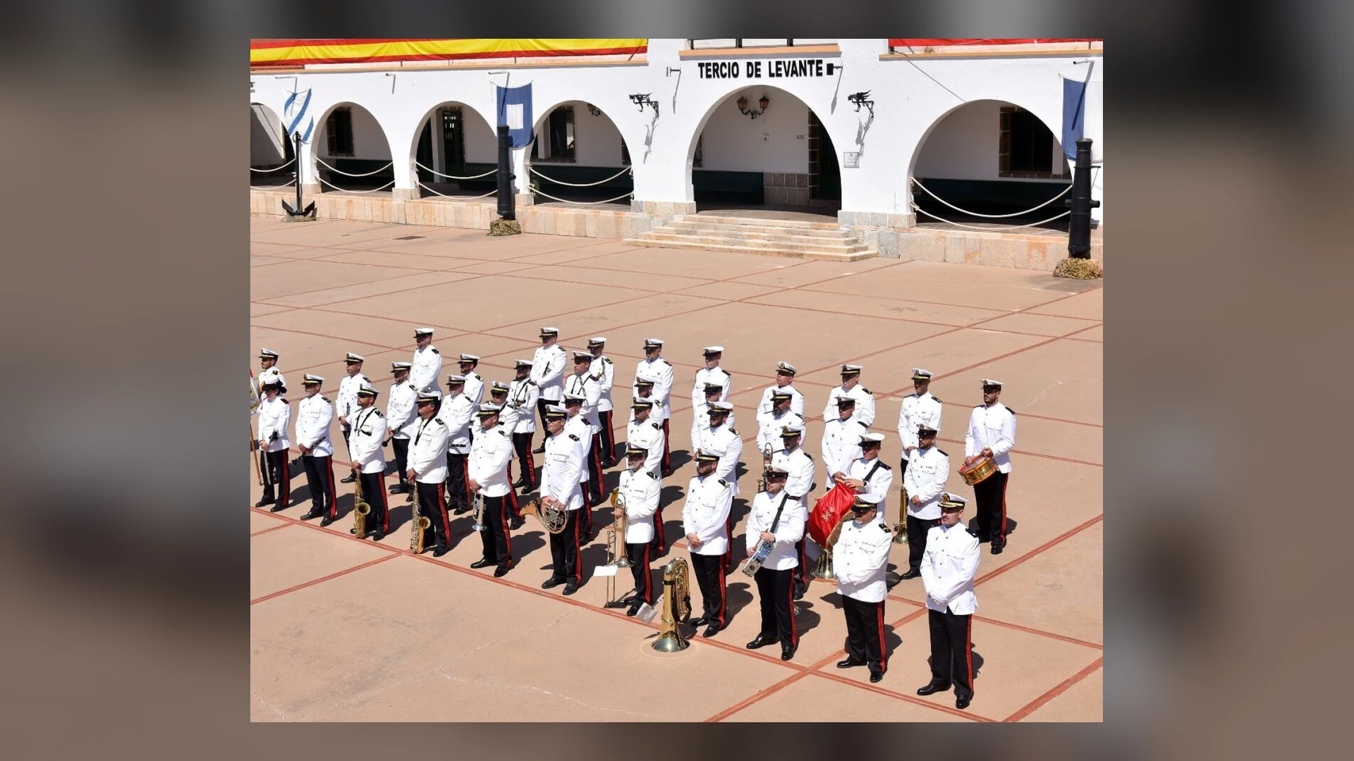 Cartel del Concierto Día de la Fiesta Nacional con una fotografía de la Unidad de Música del Tercio de Levante formada en un patio, con uniforme blanco e instrumentos de viento y percusión, frente a un edificio blanco con arcos y el rótulo Tercio de Levante